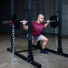 A man in a maroon t-shirt and gray shorts performs a squat on a black power rack with a barbell and weight plates in a gym setting.