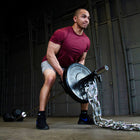 A man in a maroon t-shirt and gray shorts lifts a barbell loaded with a heavy steel chain in a dark industrial gym setting.