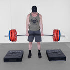 A man in a gym setting lifts a loaded barbell while standing between two black foam pads branded 'Body Solid'. He is wearing a black cap, an olive green tank top with a 'Body Solid' graphic, gray shorts, and black sneakers.