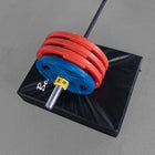 A black padded weight stack or safety pad on a gray floor, with stacked red and blue weight plates on a barbell resting on top.