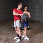 A man in a red t-shirt and gray shorts holds a black medicine ball next to a woman in a white tank top and blue shorts inside a gym with a concrete floor and gray brick walls.