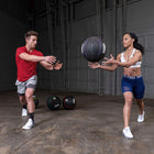 A man and a woman are exercising in a warehouse gym setting, with two medicine balls on the floor between them.