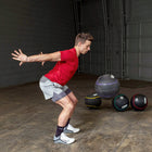 A man in a red t-shirt and gray shorts performing a squat in a gym, with medicine balls on the floor nearby.