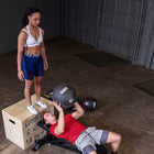 A man in a red shirt lies on a weight bench lifting a black medicine ball while a woman stands on a wooden box nearby in a garage-style gym setting.