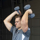 A man in a gray athletic shirt performing a shoulder press exercise with two gray hex dumbbells against a dark garage door background.