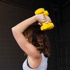 A woman in a gym lifting a bright yellow hexagonal dumbbell overhead, shown from behind. She has brown hair and is wearing a light gray tank top against a dark gray brick wall background.