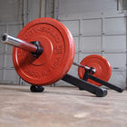 A red and black bodyweight barbell stand holding a barbell with red weight plates, photographed in a garage setting with a concrete floor and metal door in the background.
