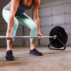 A person in mint green leggings and a gray tank top lifts a barbell with a black weight plate on a stand in a garage gym setting.