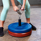 A person in mint green leggings and black sneakers uses a metal hand grip tool to lift a weight stack. The stack features a blue top weight plate with embossed text, a red bottom weight plate, and a black central post, set on a concrete floor.
