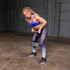 A woman in a blue sports bra and gray-to-purple gradient leggings performs a bent-over barbell carry on a garage floor in front of gray garage doors.