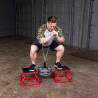 A man in athletic wear performs a squat on a red step stool while holding a chain attached to a stack of weight plates, with a second red step stool beneath him in a warehouse setting.
