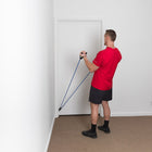 A man in a red t-shirt and black shorts stands in a room, using a blue resistance band with door handles attached to a white door.
