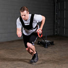 A man in athletic wear wearing a black chest support harness with an attached red handlebar grip, running in a garage setting with a weight sled in the background.