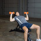 A man lying on a black adjustable weight bench in a garage setting, holding orange dumbbells overhead.