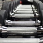 Close-up of a row of adjustable dumbbells with textured silver handles and black weight plates in a gym setting.