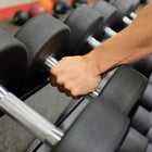 A close-up of a person's hand gripping a black and silver dumbbell in a gym setting, with other weights blurred in the background.