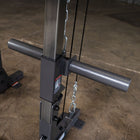 Close-up of a dark gray cable pulley weight stack machine component, showing the vertical post, horizontal handlebar, cable system, and metal chain on a concrete floor.