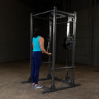 A woman stands in front of a dark power rack in a warehouse setting, holding a bar while wearing a bright turquoise tank top, navy leggings, and gray sneakers. The rack has a loaded weight plate on the side.