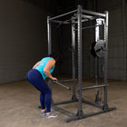 A woman in a turquoise tank top and navy leggings uses a cable pulley exercise machine inside a dark gym with concrete floors and brick walls.