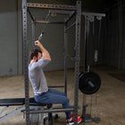 A man seated on a cable pulley strength trainee rig in a gym setting, performing a lat pulldown exercise. The rig features a black steel frame, weight stack, and cable system.