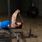 A man in a blue shirt lies on a weight bench inside a power rack, pulling a chain attached to a hanging weight plate. The setting appears to be a home gym with concrete floors and dark walls.