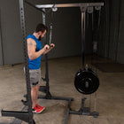 A man in a blue sleeveless shirt and gray shorts uses a cable pulley weight machine with a black weight plate in a gym setting.