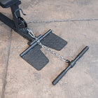 Black metal gym chain link nunchucks with textured diamond-plate foot rests, shown attached to a weight bench on a concrete floor.