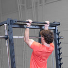 A man in an orange t-shirt is shown from behind, pulling a barbell on a black power rack with safety grips in a garage setting.