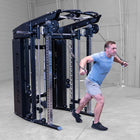 A man in a light blue t-shirt and gray shorts uses a cable machine attachment in a gym setting. The cable machine is black and silver.