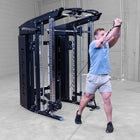 A man in a gym using a dual cable pulley system attached to a curved weight bench, wearing a light blue t-shirt, gray shorts, and black sneakers.