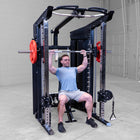 A man in a light blue t-shirt and gray shorts sits on a black and silver multi-functional strength training gym machine, performing a barbell bench press with red weight plates in a clean indoor gym setting.