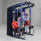 A man in a light blue t-shirt and gray shorts is using a dual-post cable pulley gym machine with red weight plates in a warehouse gym setting.