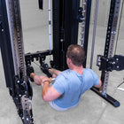A man in a light blue t-shirt sits on the floor using a cable pulley exercise machine in a gym setting.