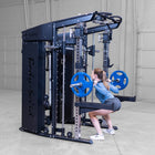 A woman performing a squat on a barbell rack attachment within a large black dual-functional gym machine, set in a concrete-floored warehouse gym environment.