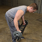 A man in a gray sleeveless shirt and sweatpants bends over in a garage while using a black metal step ladder to lift a barbell with weight plates.