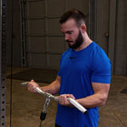 A man in a blue athletic t-shirt is shown in a gym setting, holding a silver metal cable pulley handlebar attachment.