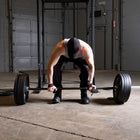 A man in a gym setting, wearing a black tank top, black pants, and a black cap, preparing to lift a barbell loaded with weights in a garage-style workout space.