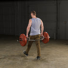 A man viewed from behind in a gym, carrying a barbell with red weight plates. 