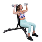 A woman sits on a black adjustable weight bench holding dumbbells overhead, wearing a pastel sports bra and mint green leggings against a plain white background.