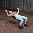 A woman in a purple and mint green sports bra and matching leggings performs a bench press on a weight bench with dumbbells in a gym setting.