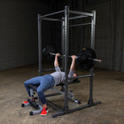 A person bench pressing with a barbell on a weight bench inside a power rack, shown in a gym setting with concrete floors and a garage door in the background.