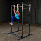 A woman in a bright blue tank top and dark leggings hangs from a pull-up bar on a black metal power rack inside a warehouse gym.