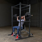 A man in a gym setting sits on a power rack bench, performing a barbell bench press. The power rack is black steel with weight plates on a barbell.
