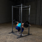 A woman in a squat position holding a barbell on a power rack inside a dark gym with concrete floors and walls.
