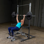 A woman in a teal tank top and blue leggings is bench pressing a loaded barbell on a silver and black power rack in a warehouse gym setting.