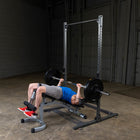 A man in a blue shirt and gray shorts lies on a weight bench pressing a barbell on a power rack inside a warehouse gym.