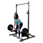 A woman in a teal tank top and blue leggings lifts a barbell loaded with black weight plates from a black power rack, shown against a plain white background.