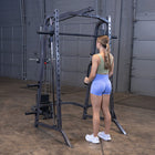 A woman in athletic wear stands in front of a dark gray cable pulley gym machine inside a warehouse or gym setting with a concrete floor and metal garage doors.