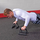 A man in a white long-sleeve athletic shirt and black-and-white patterned shorts performs pushups using black adjustable workout support bars on a dark asphalt floor in front of a gray brick wall.