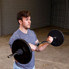 A man in a gray heather t-shirt performing a barbell curl in a garage setting with concrete floors and gray brick walls.